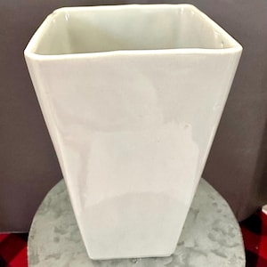 May include: A white, square-shaped vase with a slightly tapered design. The ceramic vase has a glossy finish and sits on a round, metallic surface. The vase is empty and ready to be filled with flowers or other decorative items.