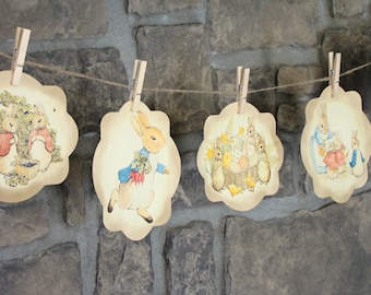 May include: A garland of scalloped-edge paper cutouts featuring illustrations of rabbits in various scenes. The cutouts are held by wooden clothespins on a twine string, with a stone wall in the background. The illustrations have a vintage, storybook aesthetic.