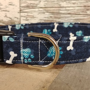 May include: A blue dog collar with a white bone and paw print pattern. The collar has a black buckle and a silver D-ring.