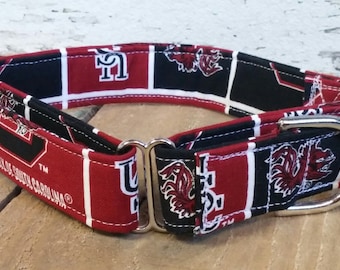 May include: A dog collar featuring a repeating pattern of red, black, and white squares with the University of South Carolina logo and text. The collar has a metal buckle and D-ring.