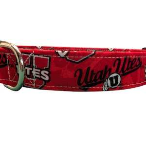 May include: Red dog collar featuring the words "Utah Utes" in black script and white block letters. The collar has a black plastic buckle and a silver metal D-ring for leash attachment. The pattern includes the Utes logo and other team-related graphics.