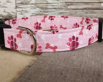 May include: Pink dog collar with a paw print and bone pattern. The collar has a black buckle and a silver D-ring.