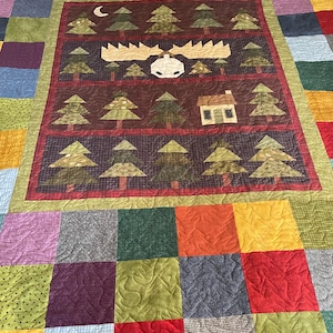 May include: A patchwork quilt with a central panel featuring a moose head, trees, and a small house. The quilt has a variety of colorful squares and a border. The color palette includes reds, greens, browns, and yellows.