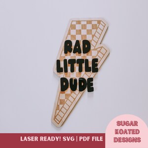 May include: Wooden lightning bolt-shaped sign with a checkerboard pattern and the words "RAD LITTLE DUDE" in black letters. The sign also includes the text "LASER READY! SVG | PDF FILE" and "SUGAR KOATED DESIGNS".