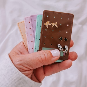 May include: A brown earring display card with gold bow earrings, black heart earrings, and checkered hoop earrings. The display is held with several pastel-colored earring display cards.