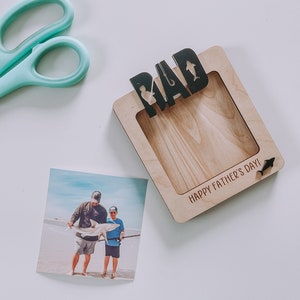 Może przedstawiać: Drewniana ramka na zdjęcie z napisem "DAD" i sylwetką wędki i ryby. Ramka ma miejsce na zdjęcie i napis "Happy Father's Day!" na dole.