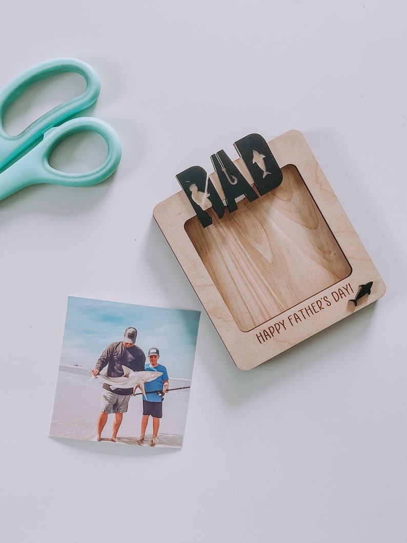 Może przedstawiać: Drewniana ramka na zdjęcie z napisem "DAD" i sylwetką wędki i ryby. Ramka ma miejsce na zdjęcie i napis "Happy Father's Day!" na dole.