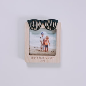 K&ouml;nnte beinhalten: Ein Holz-Bilderrahmen mit einem schwarzen Ausschnitt von einer Sonnenbrille, auf dem oben "Rad Dad" steht. Der Rahmen enth&auml;lt ein Foto von einem Vater und seinem Sohn am Strand mit einem Hund. Der Rahmen tr&auml;gt auch den Text "Happy Father's Day! 2024" unten.