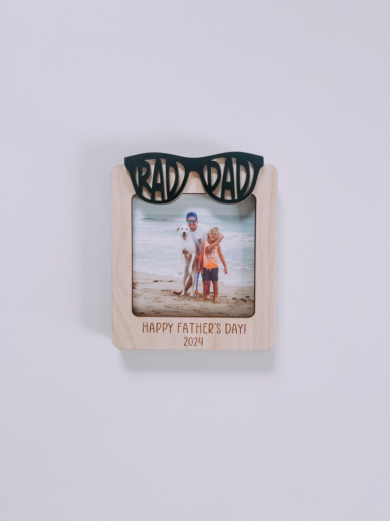 K&ouml;nnte beinhalten: Ein Holz-Bilderrahmen mit einem schwarzen Ausschnitt von einer Sonnenbrille, auf dem oben "Rad Dad" steht. Der Rahmen enth&auml;lt ein Foto von einem Vater und seinem Sohn am Strand mit einem Hund. Der Rahmen tr&auml;gt auch den Text "Happy Father's Day! 2024" unten.