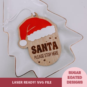 Op de afbeelding: Houten kerstversiering met een rode kerstmanmuts en de tekst "Santa Please Stop Here".
