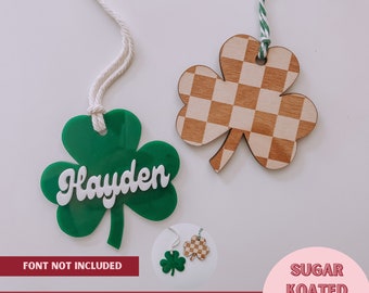May include: Two laser-cut shamrock shapes, one green with the name "Hayden" in white lettering, and one wood with a brown and white checkered pattern. The text "FONT NOT INCLUDED" and "LASER READY! SVG FILE" are at the bottom of the image. The text "SUGAR KOATED DESIGNS" is in the lower right corner.