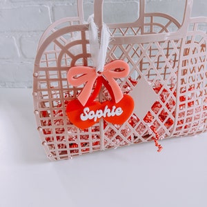 May include: Pink plastic tote bag with a red heart-shaped tag that says "Sophie" and a pink bow.