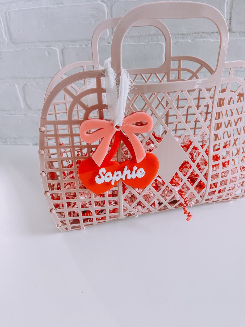 May include: Pink plastic tote bag with a red heart-shaped tag that says "Sophie" and a pink bow.