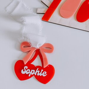 May include: Red and pink cherry-shaped acrylic ornament with the name "Sophie" written in white. The ornament has a pink bow.