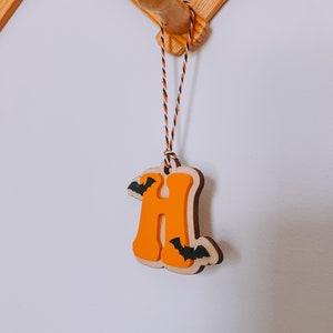 May include: Orange wooden letter H with black bats hanging from a string. The letter is hanging from a wooden hook.