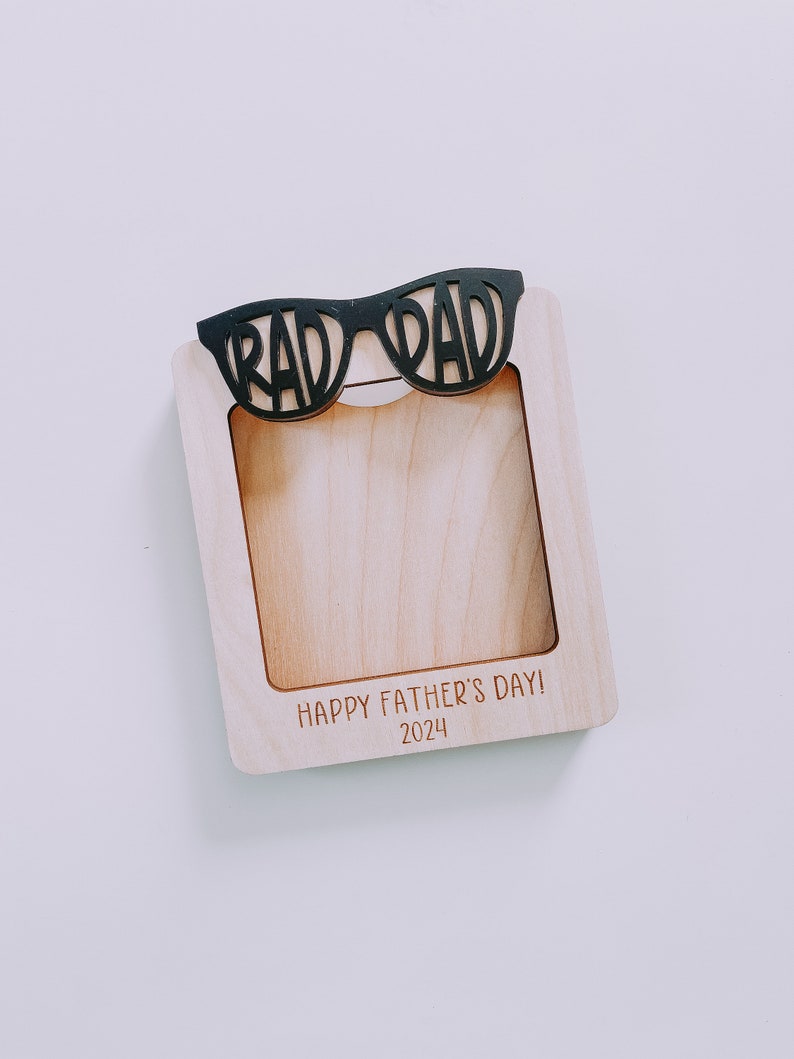 K&ouml;nnte beinhalten: Ein Holzbildrahmen mit einem Ausschnitt von einer Sonnenbrille, auf dem oben "Rad Dad" steht. Der Rahmen hat den Text "Happy Father's Day! 2024" unten eingraviert.