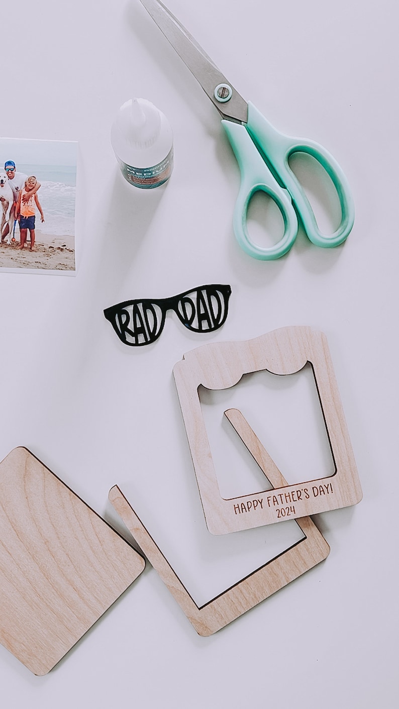 K&ouml;nnte beinhalten: Ein DIY-Bastelset zum Vatertag. Das Set enth&auml;lt einen Holzrahmen mit dem Text "Happy Father's Day! 2024", eine schwarze Papier-Sonnenbrille mit dem ausgeschnittenen Wort "RAD DAD" und ein Foto einer Familie am Strand.