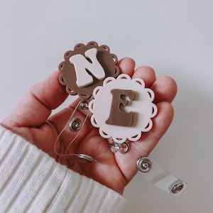 K&ouml;nnte beinhalten: Zwei runde Ausweishalter mit gewelltem Rand. Ein Halter ist braun mit einem cremefarbenen Buchstaben N, der andere ist cremefarben mit einem braunen Buchstaben E. Beide haben durchsichtige Kunststoffb&auml;nder mit Metalldruckkn&ouml;pfen zur Befestigung.