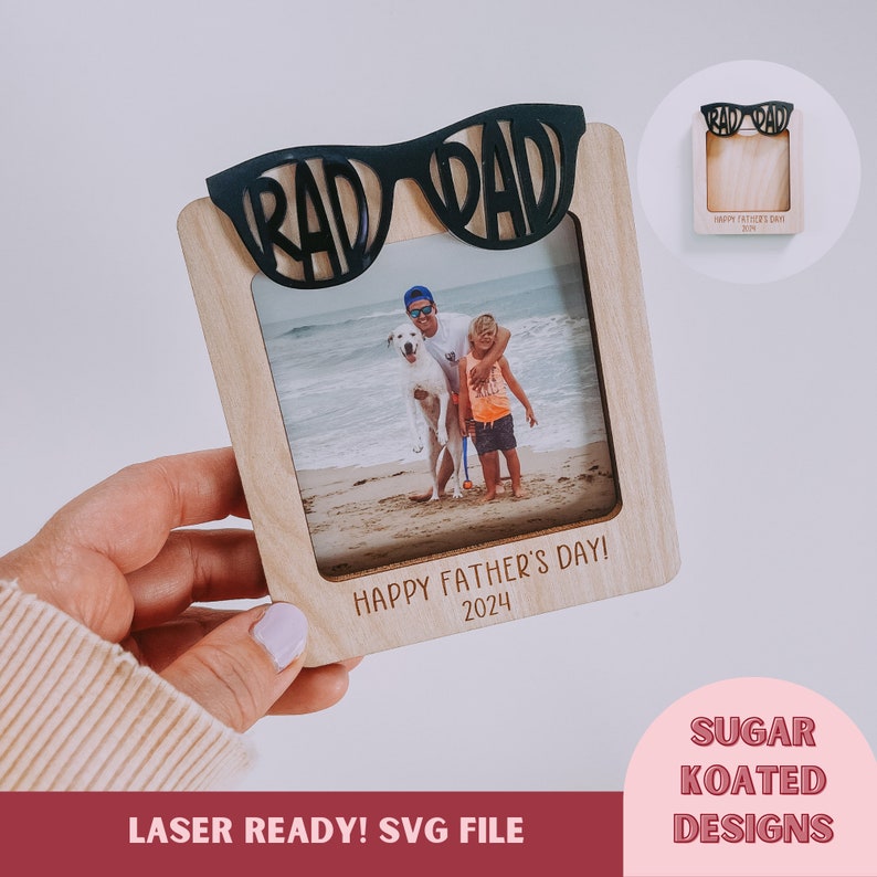 K&ouml;nnte beinhalten: Ein Holzbildrahmen mit einem schwarzen Ausschnitt von einer Sonnenbrille, auf dem oben "RAD DAD" steht. Der Rahmen enth&auml;lt ein Foto von einem Vater und seinem Sohn am Strand mit einem Hund. Der Text "HAPPY FATHER'S DAY! 2024" ist auf den Rahmen gedruckt.