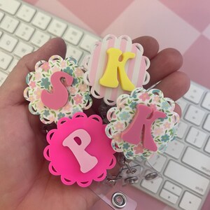 K&ouml;nnte beinhalten: Drei runde Ausweishalter mit gewelltem Rand. Einer hat ein rosa Blumenmuster mit einem rosa Buchstaben S, einer hat rosa-wei&szlig;e vertikale Streifen mit einem gelben Buchstaben K und einer hat einen kr&auml;ftig rosa Hintergrund mit einem hellrosa Buchstaben P.