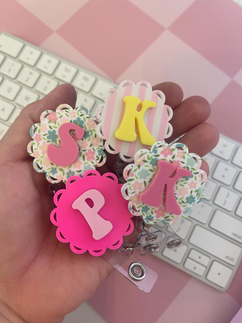 K&ouml;nnte beinhalten: Drei runde Ausweishalter mit gewelltem Rand. Einer hat ein rosa Blumenmuster mit einem rosa Buchstaben S, einer hat rosa-wei&szlig;e vertikale Streifen mit einem gelben Buchstaben K und einer hat einen kr&auml;ftig rosa Hintergrund mit einem hellrosa Buchstaben P.