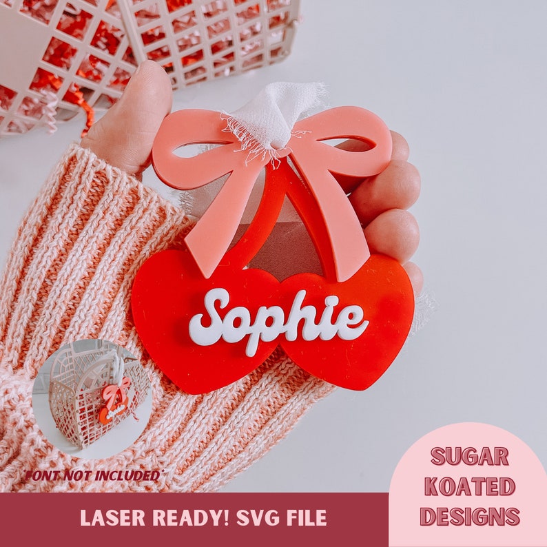 May include: Red and pink cherry-shaped ornament with the name "Sophie" written in white. The ornament is laser-cut and ready to be personalized.  The ornament is held in a hand wearing a pink knitted sweater.