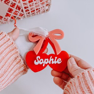 May include: Red and pink cherry-shaped acrylic ornament with the name "Sophie" written in white letters.