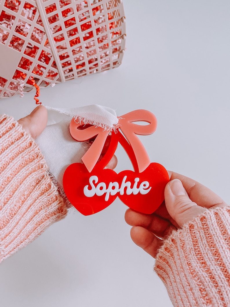 May include: Red and pink cherry-shaped acrylic ornament with the name "Sophie" written in white letters.