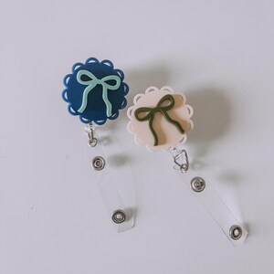 Op de afbeelding: Twee badgehaspels met geschulpte randen en strikaccenten. E&eacute;n haspel is marineblauw met een mintgroene strik, de andere lichtroze met een saliegroene strik. Elke haspel heeft een doorzichtige plastic band en een metalen clip.