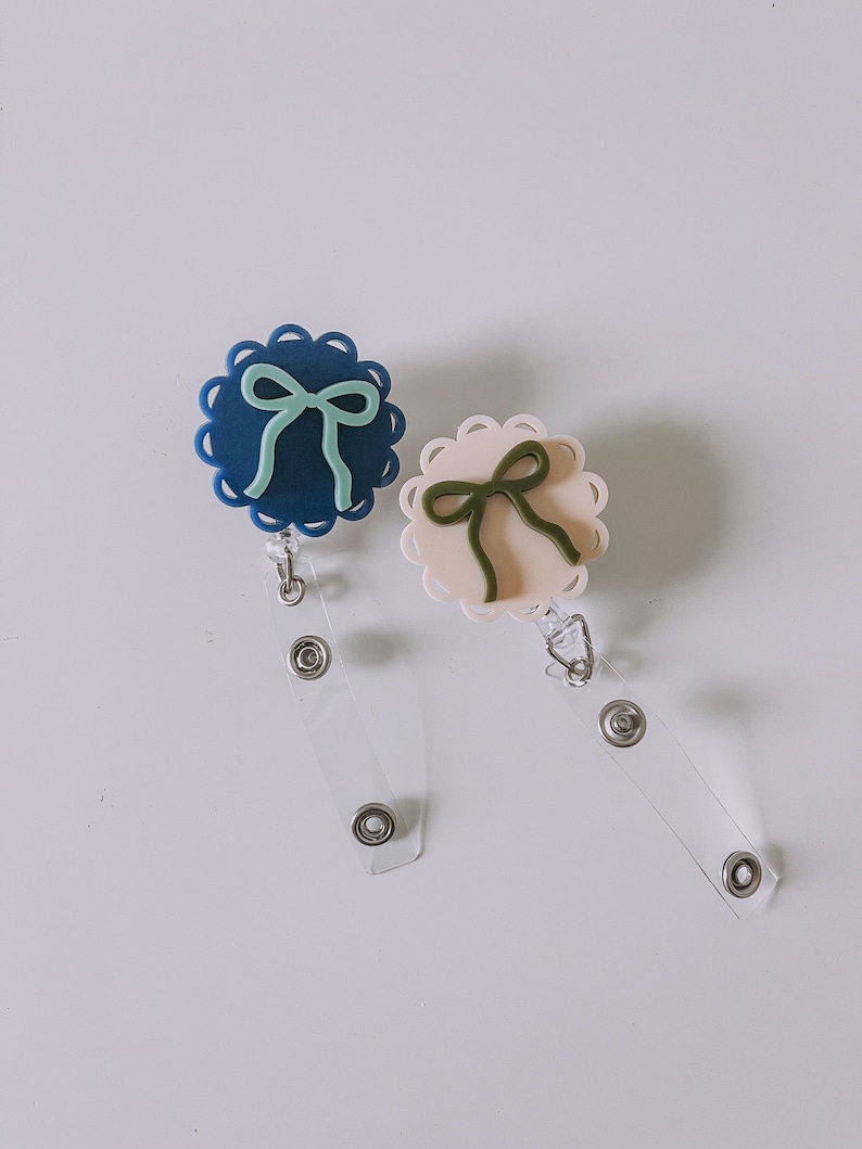Op de afbeelding: Twee badgehaspels met geschulpte randen en strikaccenten. E&eacute;n haspel is marineblauw met een mintgroene strik, de andere lichtroze met een saliegroene strik. Elke haspel heeft een doorzichtige plastic band en een metalen clip.