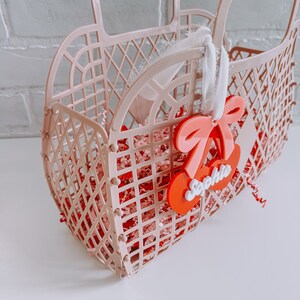 May include: A pink plastic basket with a white ribbon and a red cherry charm with the name "Sophie" on it. The basket is filled with pink and white crinkle paper.