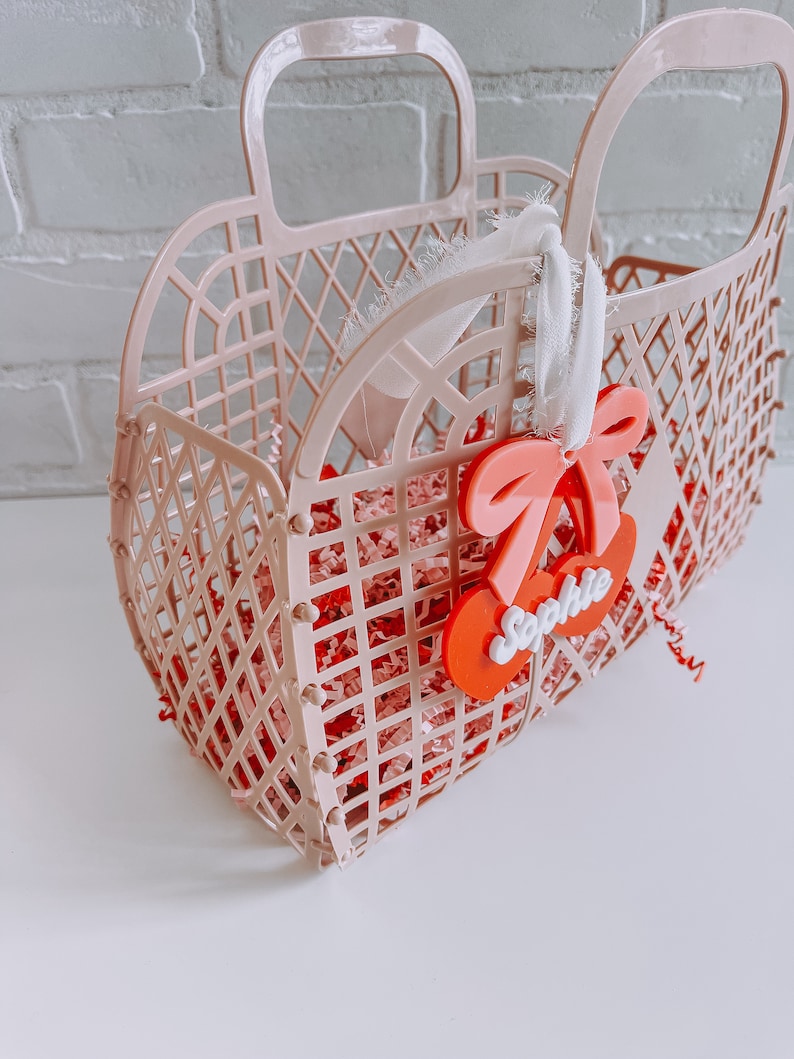 May include: A pink plastic basket with a white ribbon and a red cherry charm with the name "Sophie" on it. The basket is filled with pink and white crinkle paper.