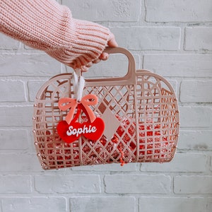 May include: A pink plastic basket with a red heart-shaped tag that says "Sophie" hanging from the handle. The basket is filled with pink and red crinkle paper.