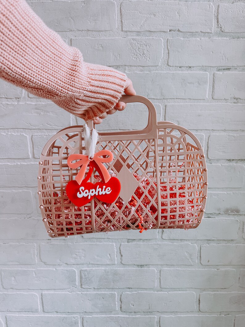 May include: A pink plastic basket with a red heart-shaped tag that says "Sophie" hanging from the handle. The basket is filled with pink and red crinkle paper.