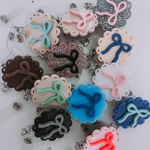 Op de afbeelding: Een verzameling badge reels met geschulpte randen en strikaccenten in verschillende kleuren, waaronder roze, blauw en bruin. Elke reel heeft een doorzichtige plastic band en een metalen clip voor bevestiging aan kleding of accessoires.
