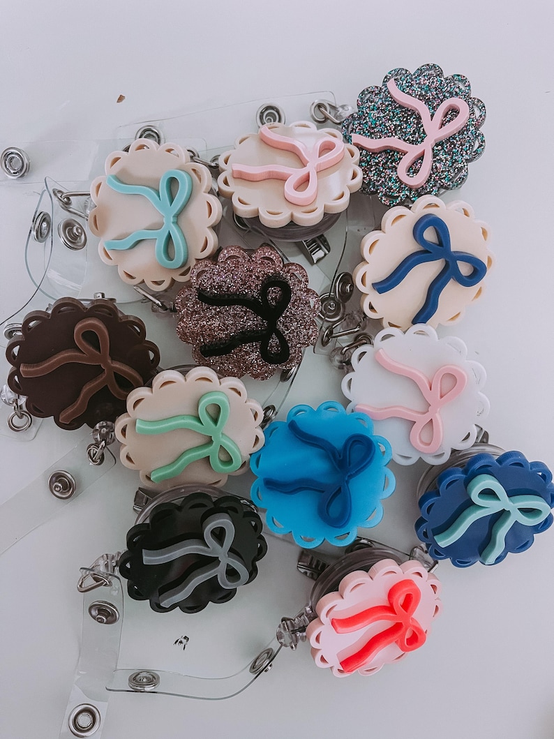 Op de afbeelding: Een verzameling badge reels met geschulpte randen en strikaccenten in verschillende kleuren, waaronder roze, blauw en bruin. Elke reel heeft een doorzichtige plastic band en een metalen clip voor bevestiging aan kleding of accessoires.