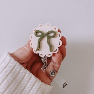 Op de afbeelding: Een badge reel met een geschulpte rand en een lichtroze midden. Een groene strik staat in het midden van de badge reel. De badge reel heeft een doorzichtige plastic band en een metalen clip.