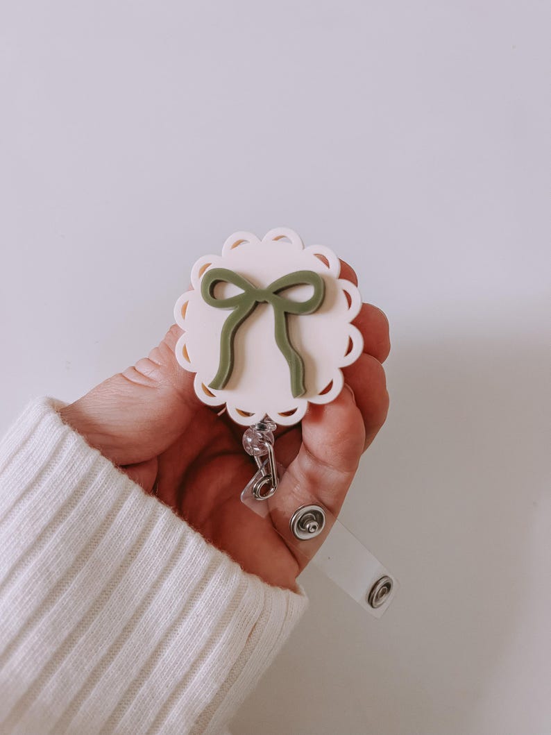 Op de afbeelding: Een badge reel met een geschulpte rand en een lichtroze midden. Een groene strik staat in het midden van de badge reel. De badge reel heeft een doorzichtige plastic band en een metalen clip.
