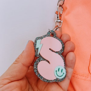 May include: Pink glitter letter S keychain with a light blue lightning bolt and a green smiley face.