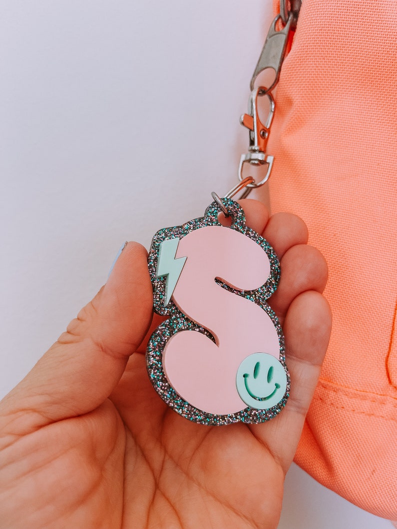 May include: Pink glitter letter S keychain with a light blue lightning bolt and a green smiley face.
