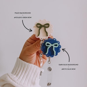Op de afbeelding: Twee badgehaspels met strikdesigns. E&eacute;n heeft een perzikachtergrond met een avocado-groene strik, en de andere heeft een donkerblauwe achtergrond met een arctische blauwe strik. Beide haspels hebben een geschulpte rand en een doorzichtig intrekbaar koord.