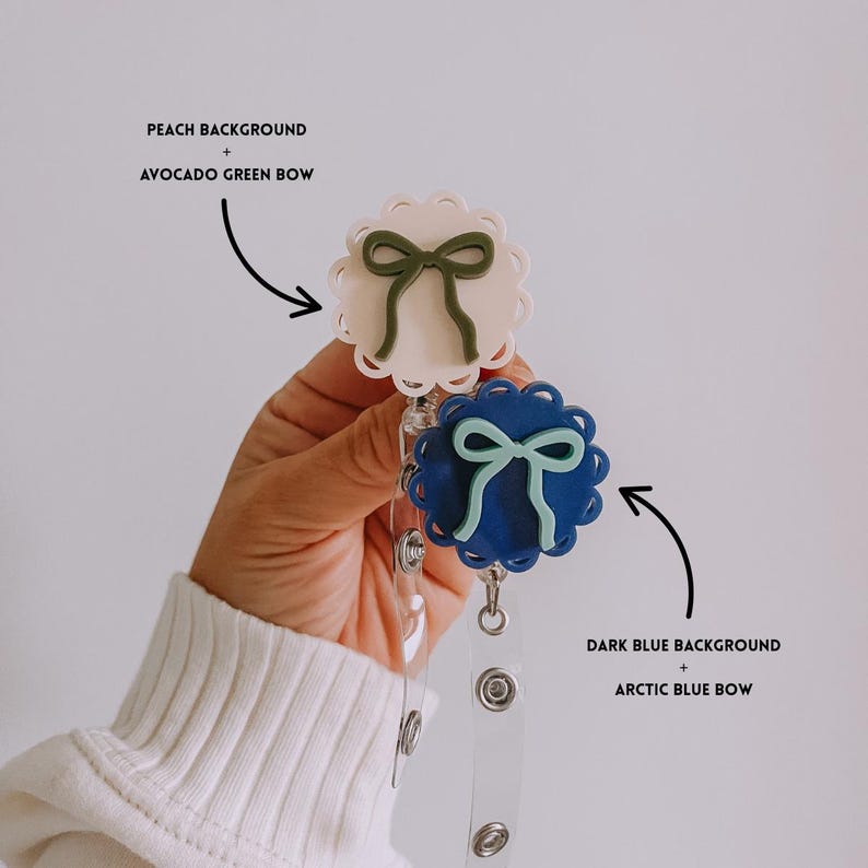 Op de afbeelding: Twee badgehaspels met strikdesigns. E&eacute;n heeft een perzikachtergrond met een avocado-groene strik, en de andere heeft een donkerblauwe achtergrond met een arctische blauwe strik. Beide haspels hebben een geschulpte rand en een doorzichtig intrekbaar koord.