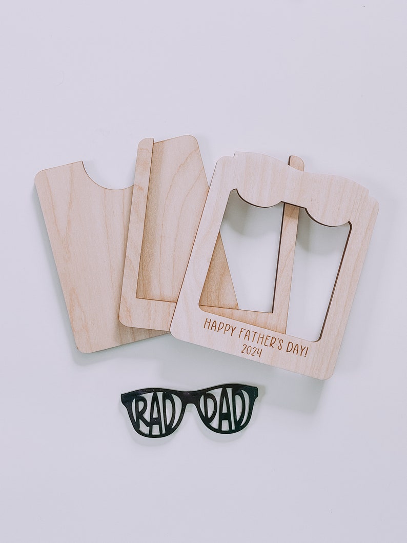 K&ouml;nnte beinhalten: Holzrahmen mit dem Text "Happy Father's Day! 2024" und einer schwarzen Sonnenbrille mit dem Text "Rad Dad" auf den Gl&auml;sern.