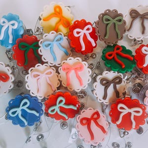 Op de afbeelding: Een verzameling kleurrijke badgehaspels met geschulpte randen en strikaccenten. De haspels hebben verschillende kleuren, waaronder rood, blauw, roze, groen en bruin, met witte strikken. Elke haspel heeft een doorzichtig plastic koord en een metalen clip.