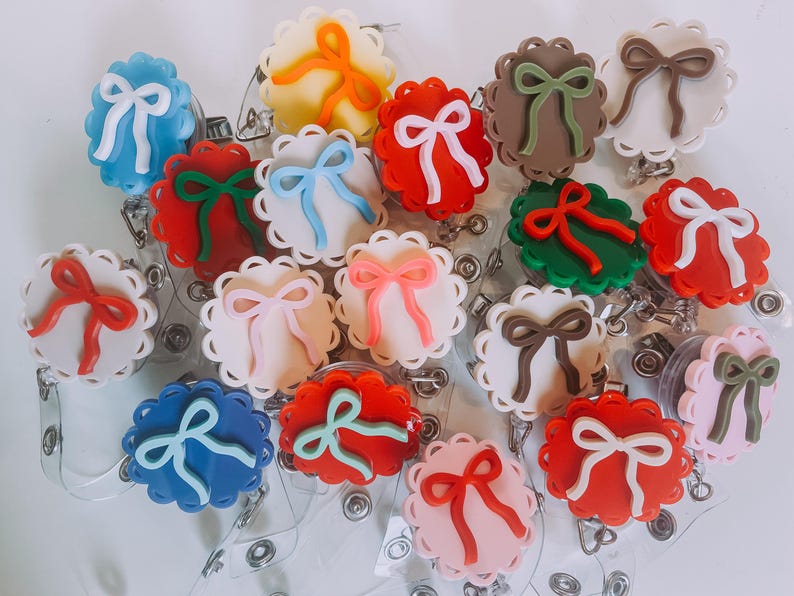 Op de afbeelding: Een verzameling kleurrijke badgehaspels met geschulpte randen en strikaccenten. De haspels hebben verschillende kleuren, waaronder rood, blauw, roze, groen en bruin, met witte strikken. Elke haspel heeft een doorzichtig plastic koord en een metalen clip.