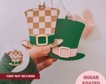 May include: Two wooden St. Patrick's Day hat ornaments. One hat has a checkerboard pattern, green band, and gold buckle. The other is green with a pink band and gold buckle. A smaller ornament with the name "Hayden" is also visible.
