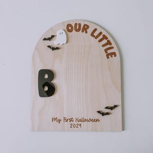 May include: A wooden sign with a rounded top that says "Our Little B" with a black letter B and a white ghost. The sign also says "My First Halloween 2024" and has black bats.