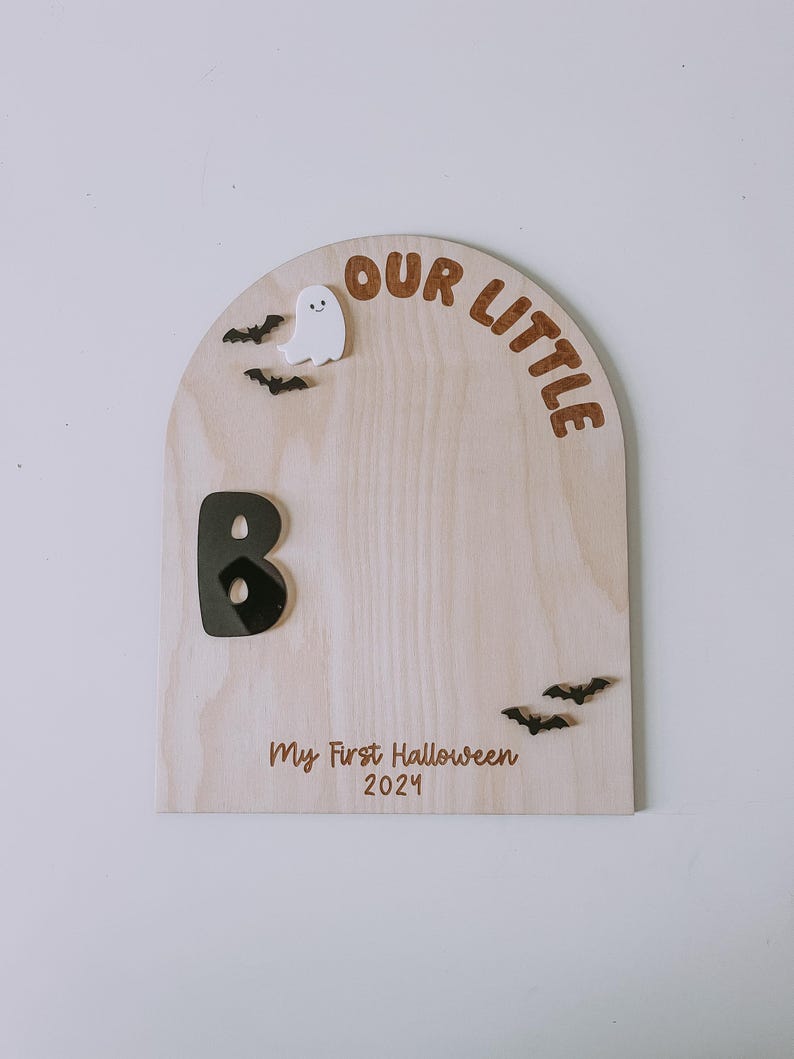 May include: A wooden sign with a rounded top that says "Our Little B" with a black letter B and a white ghost. The sign also says "My First Halloween 2024" and has black bats.