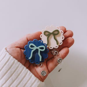 Op de afbeelding: Twee badge reels met geschulpte randen en strikaccenten. E&eacute;n reel is marineblauw met een lichtblauwe strik, de andere gebroken wit met een groene strik. Elke reel heeft een doorzichtige plastic band en een metalen clip.