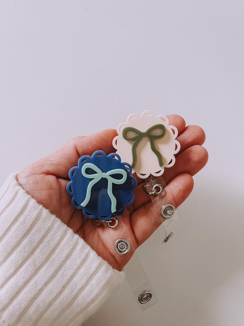 Op de afbeelding: Twee badge reels met geschulpte randen en strikaccenten. E&eacute;n reel is marineblauw met een lichtblauwe strik, de andere gebroken wit met een groene strik. Elke reel heeft een doorzichtige plastic band en een metalen clip.